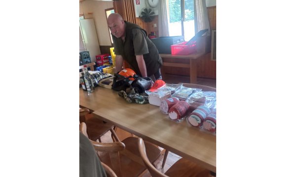 Ein Mann lehnt sich über einen Tisch, auf dem diverse Gegenstände wie Kleidung, Handschuhe und Lebensmittel liegen. Die Umgebung wirkt wie ein rustikaler, heller Innenraum mit Holzstühlen.